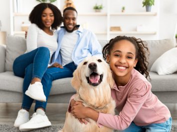 Bonding, Togetherness And Friendship. Portrait of smiling black girl hugging her happy dog in living room at home, looking at camera, cheerful parents sitting on the couch in blurred background